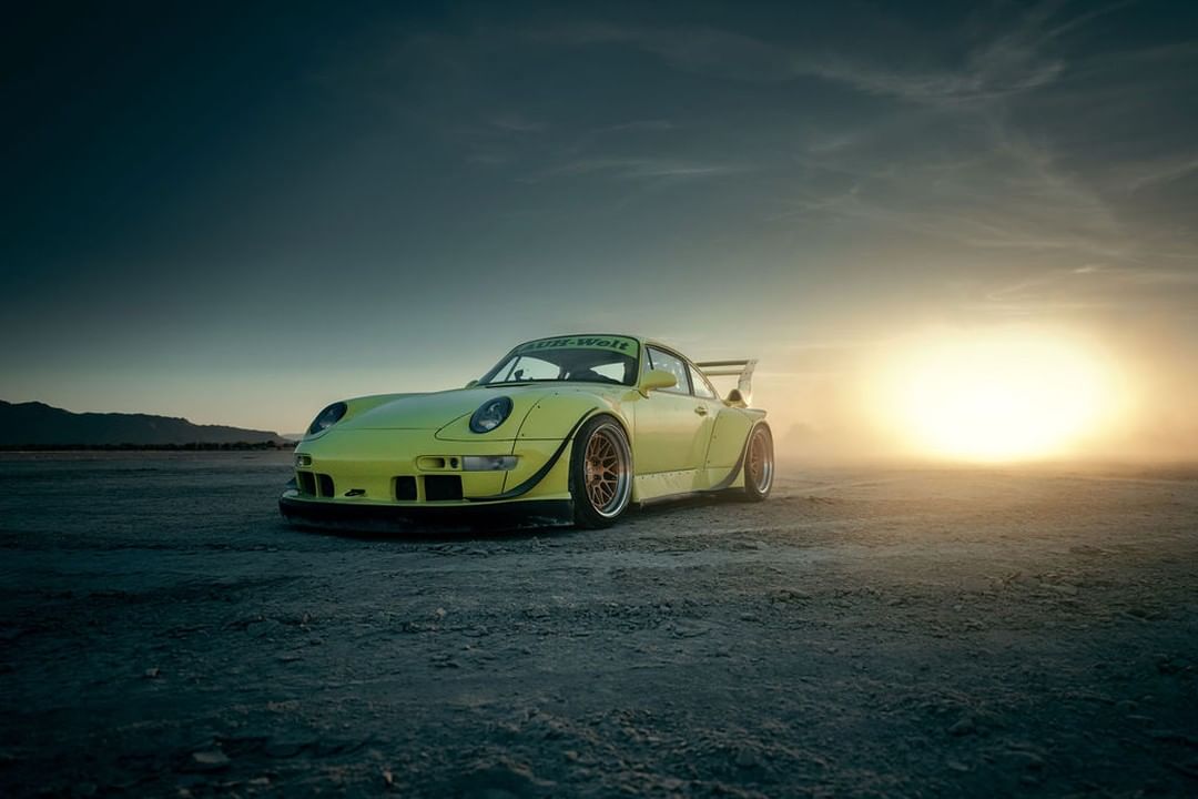 Yellow Porsche 911 parked on a vast dirt expanse under a dramatic sky with a setting sun creating a striking automotive image.
