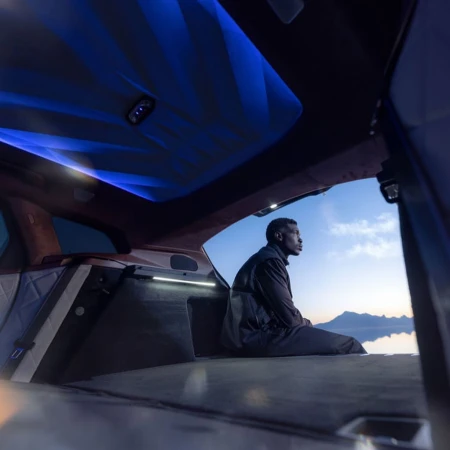 An African-American adult male sits in the open trunk of an SUV, looking out towards a vast salt flat and sky. Blue interior lights add ambiance.