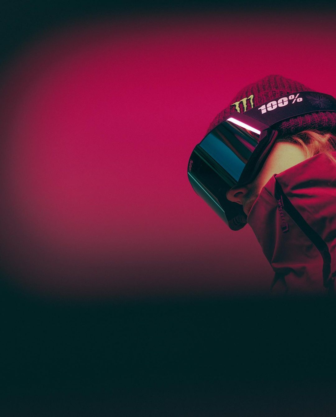 Low-angle portrait of a person wearing snow goggles and a beanie against a gradient magenta background, shot with studio lighting.