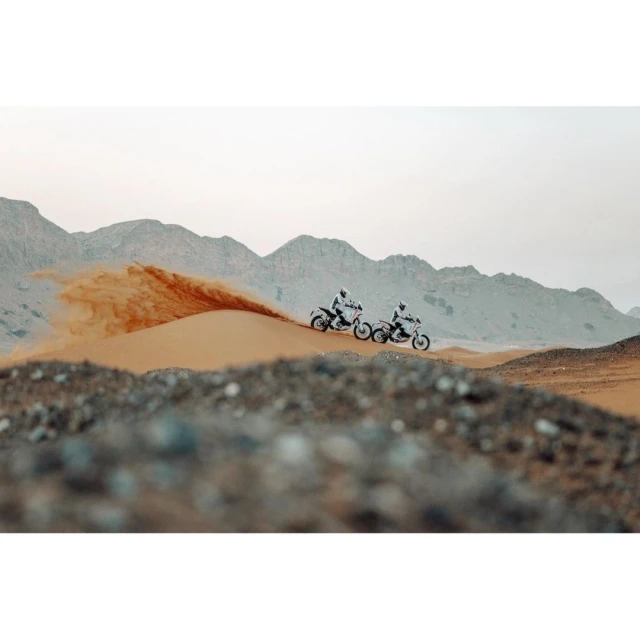 Two motorsport racers on dirt bikes speed across a desert sand dune with mountains in the background during a rally race.