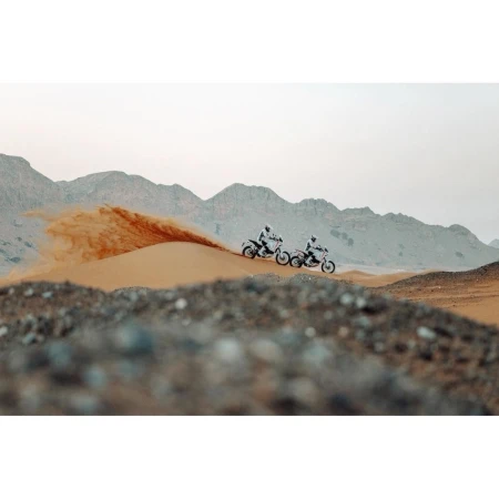 Two motorsport racers on dirt bikes speed across a desert sand dune with mountains in the background during a rally race.