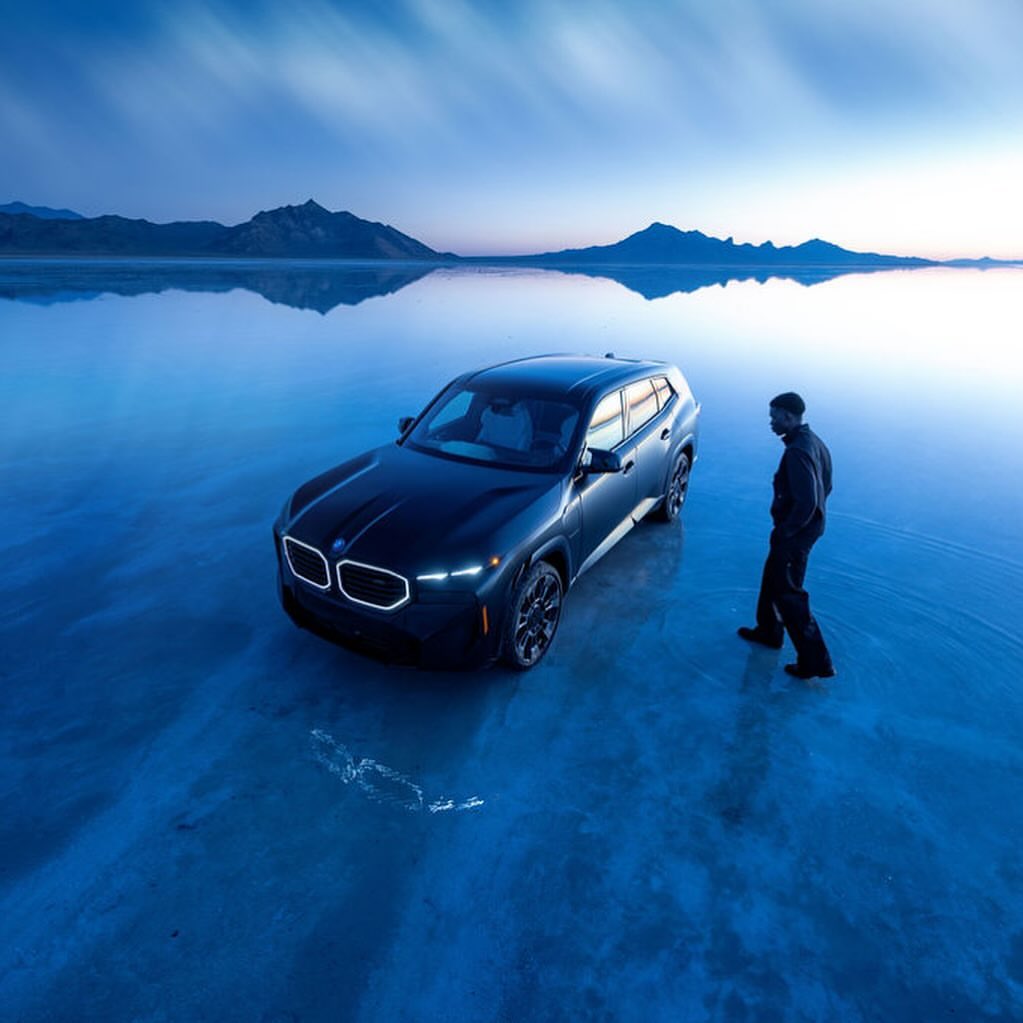 A black BMW SUV sits on a salt flat with a man walking near the vehicle under a blue sky with distant mountains in the background.