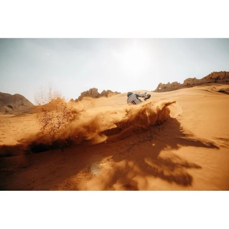 A person in a white suit sandboarding down a red-hued desert dune with sand billowing in the air, against a sunny sky backdrop.