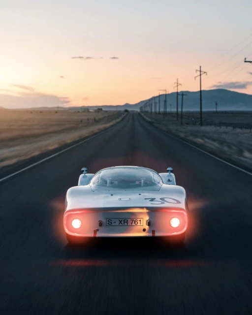 Vintage white race car drives down an empty road towards the horizon at sunset. Mountain range and telephone poles in background.