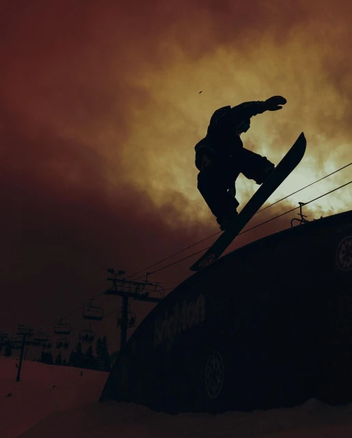 Silhouette of a snowboarder performing a trick over a snow feature, with ski lift infrastructure visible against a sunset-colored sky.