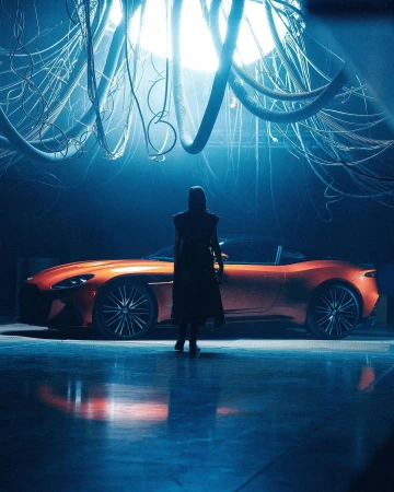 Silhouette of a person standing before a glossy orange sports car under a cluster of hanging cables in a moody studio.