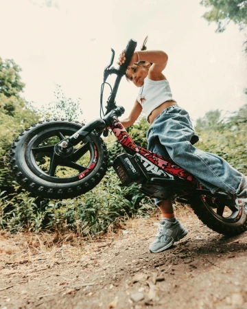 A young child rides an electric balance bike on a dirt trail, lifting the front wheel up against a natural green foliage background.