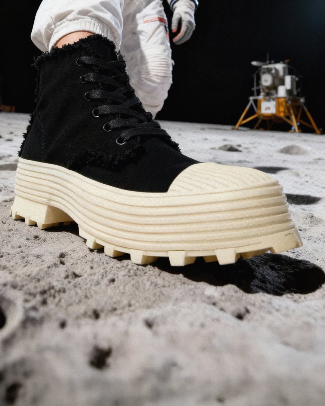 Close-up of black high-top platform boot on gray moon surface, person in space suit and lunar module in the background.