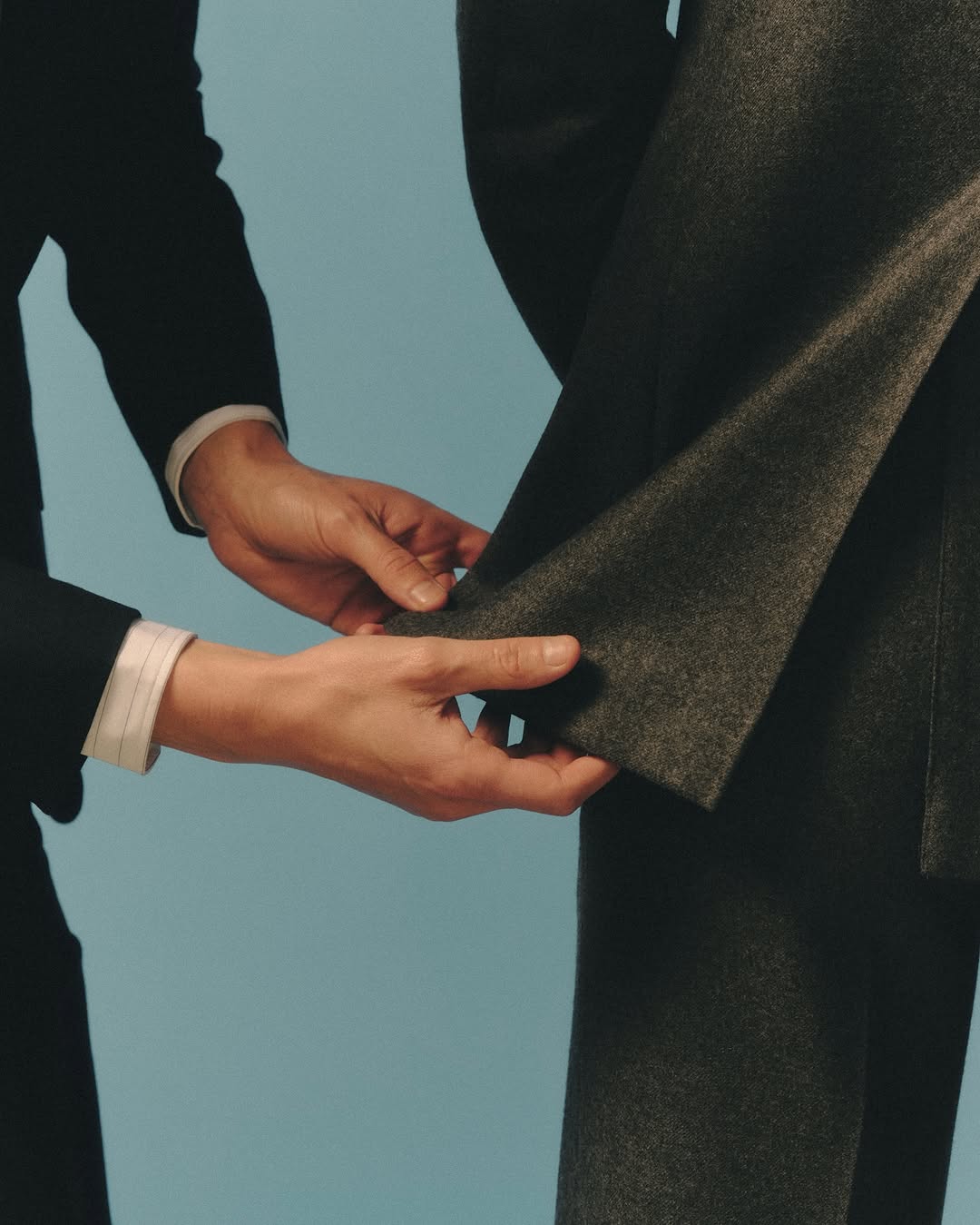 Close-up studio shot of a dark grey suit being adjusted by a person wearing a white shirt and dark jacket against a light blue backdrop.