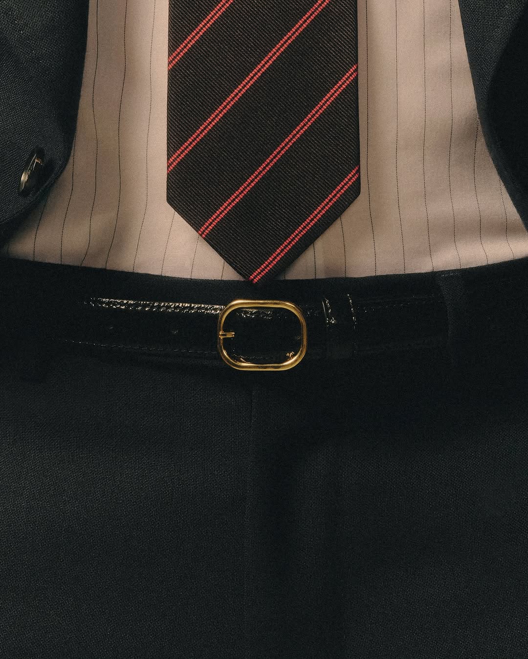 Close-up of a torso wearing a suit, striped shirt, striped tie, and black patent leather belt with gold buckle, studio lighting.