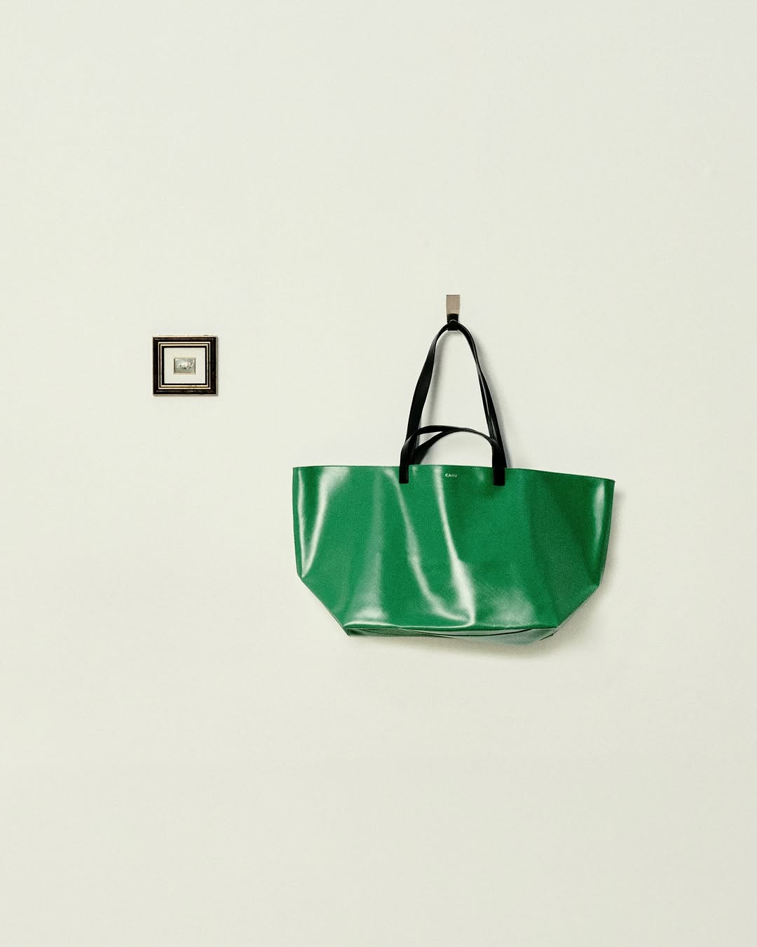 Shiny green tote bag with black handles hangs on a wall next to a small framed artwork in an indoor setting with neutral walls.