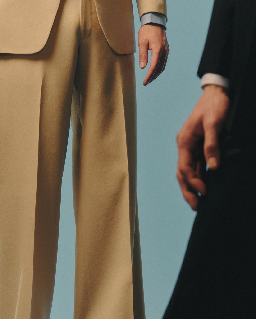 Close-up of two people wearing suits in front of light blue background. One person has a tan suit and other has black suit.