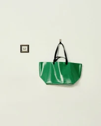 Green leather tote bag with black handles hanging on a hook against a plain light wall with a small framed picture