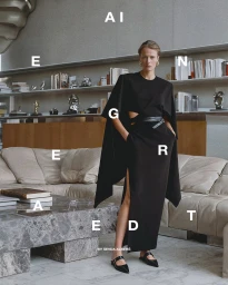 A woman in a black cape-style dress stands in a modern living room interior with bookshelves and a large marble coffee table.