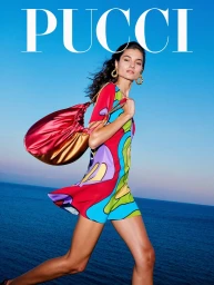 Model in colourful patterned dress carrying a red satin shoulder bag against a blue ocean background