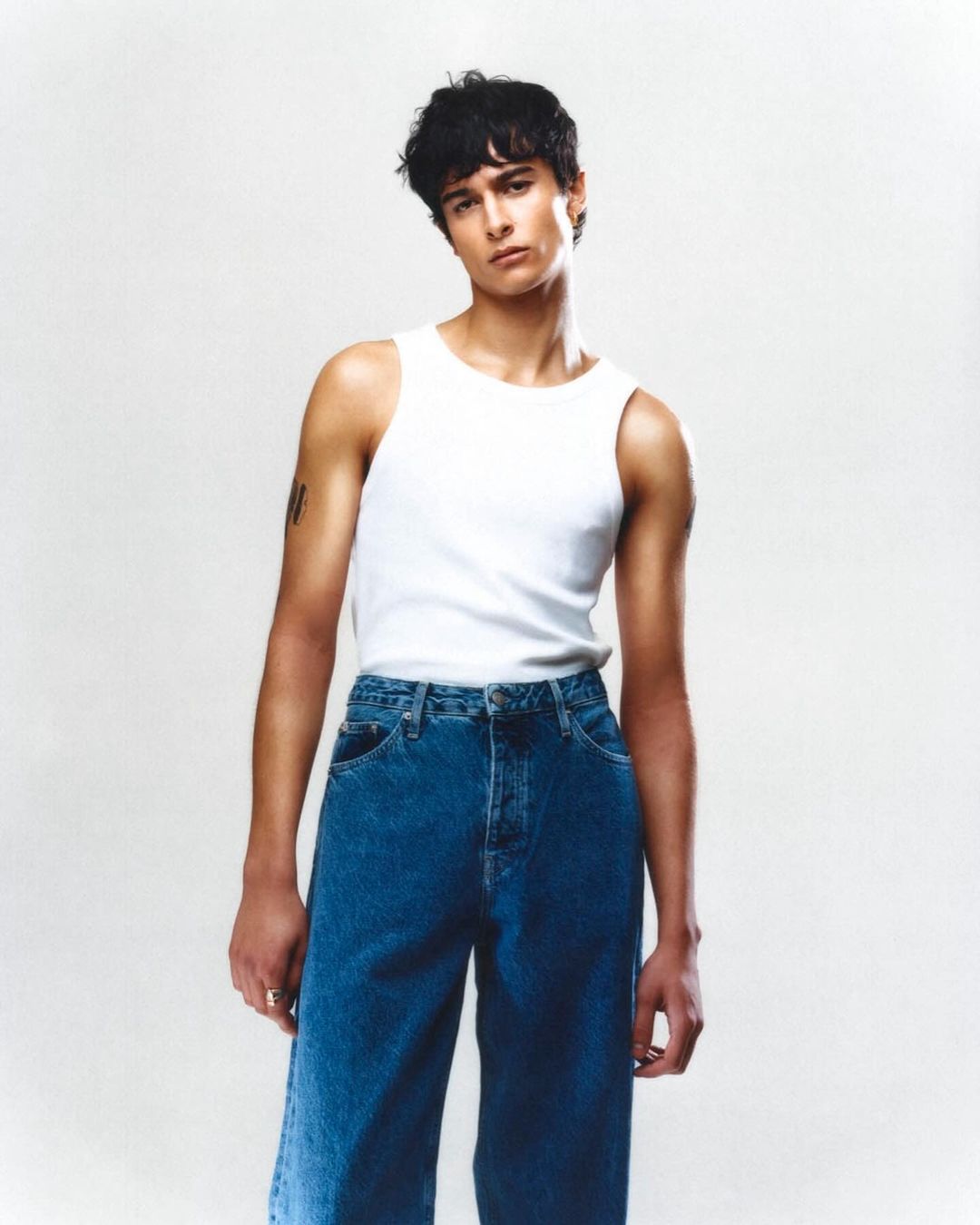 Portrait of a young adult male wearing a white tank top and blue jeans, posing in a photo studio against a clean, light-colored background.