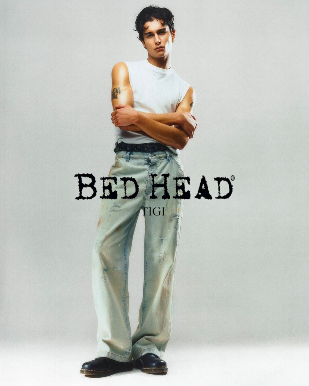 A young adult man poses in a photo studio. He is wearing light wash jeans, a white t-shirt, and black boots.
