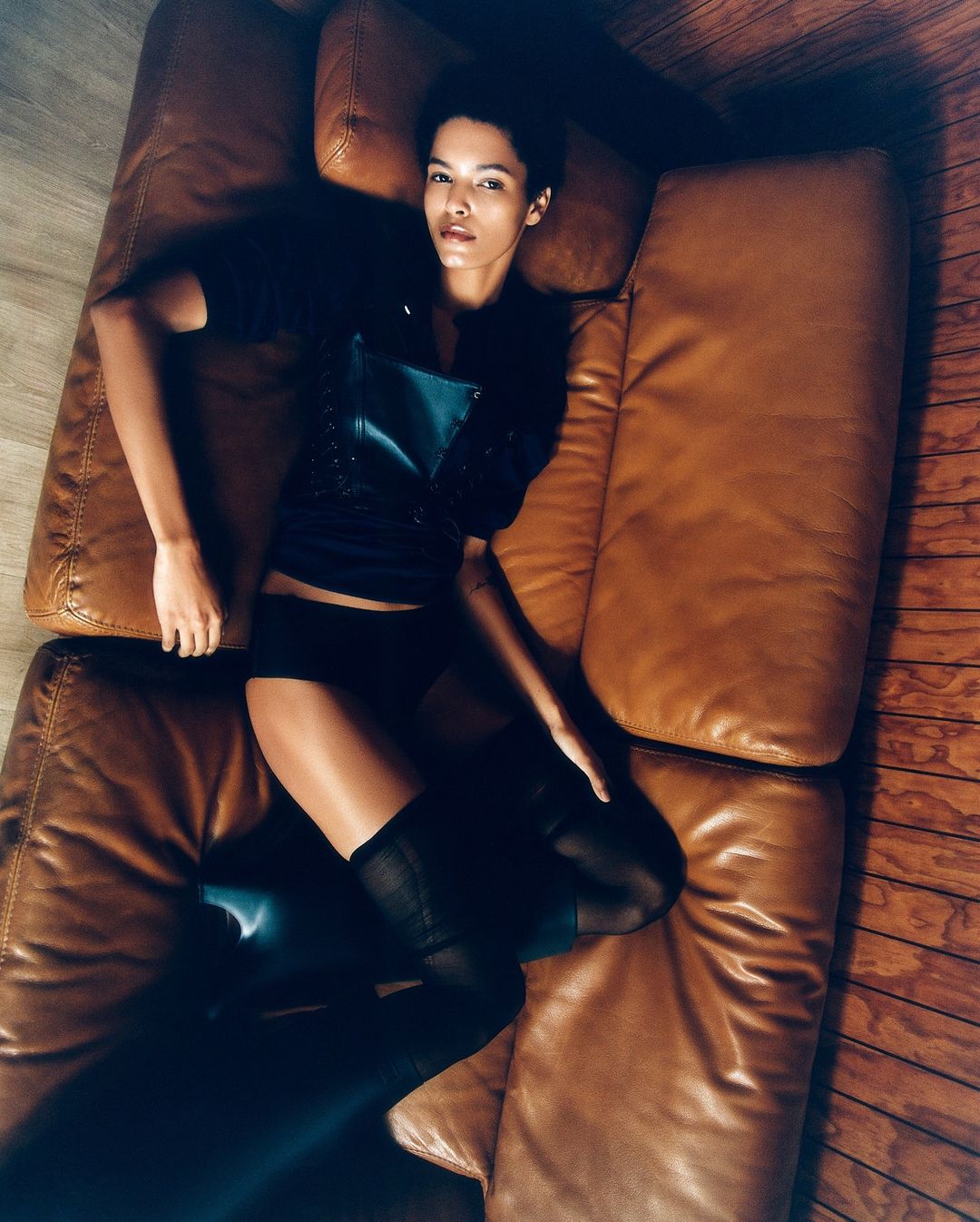 Adult woman lies on a brown leather couch wearing black top, shorts, and black stockings in a high-angle shot. Wood flooring visible.