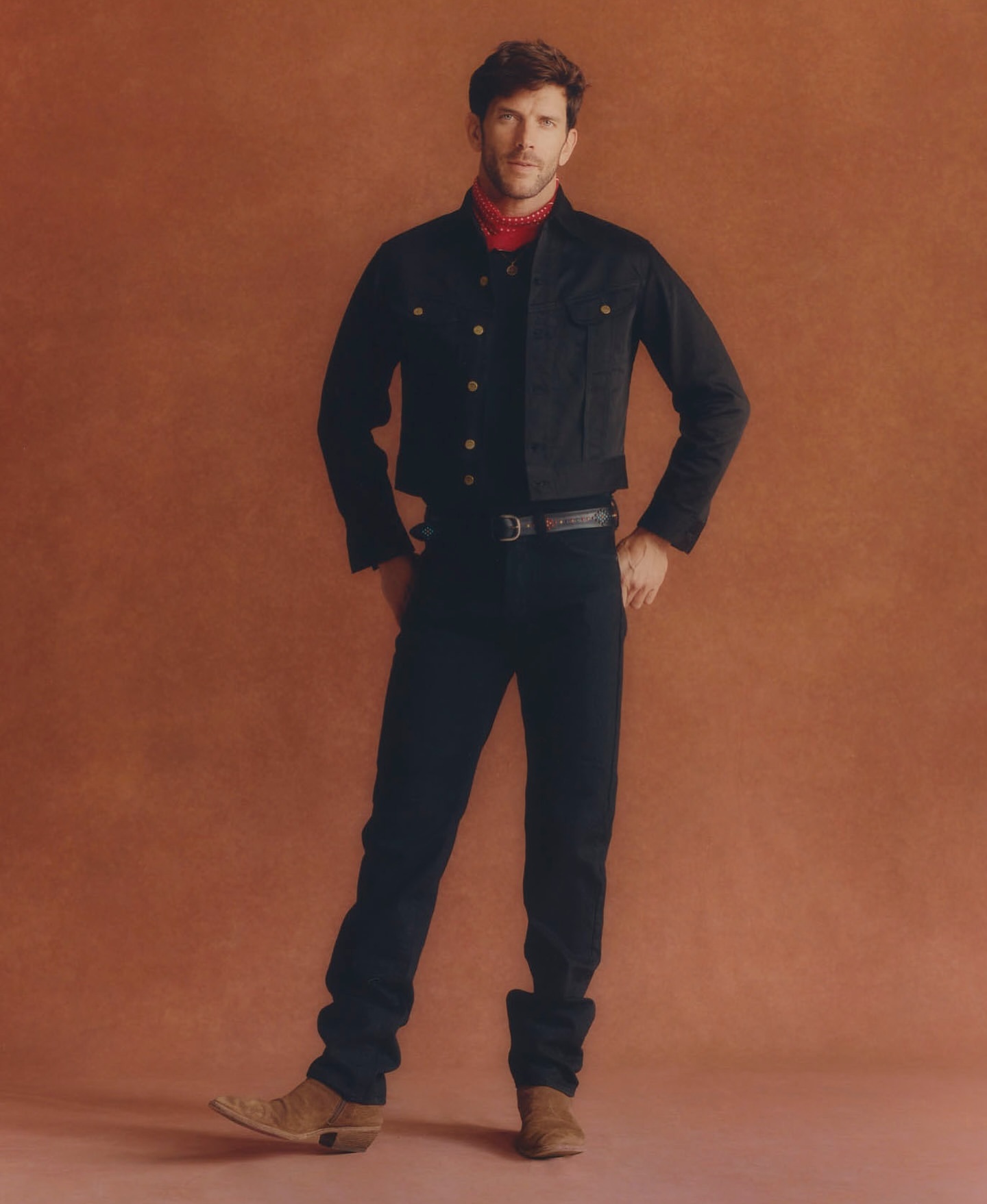 Full-length studio portrait of a man in a dark denim outfit with a red bandana, posing against a gradient brown backdrop with subtle lighting.