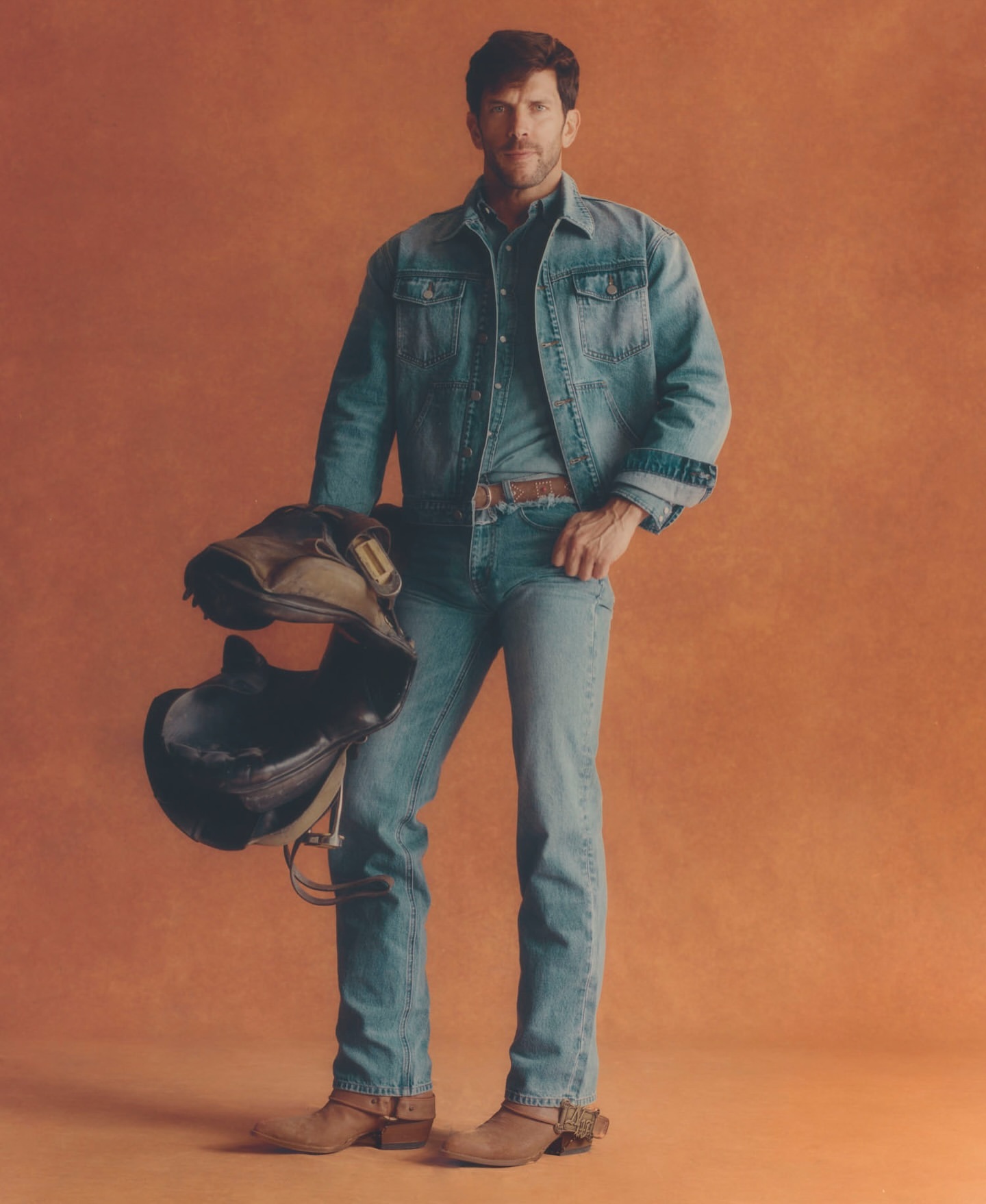 Portrait of a fair-skinned adult man in a denim jacket and jeans, holding a black helmet against an orange studio backdrop.