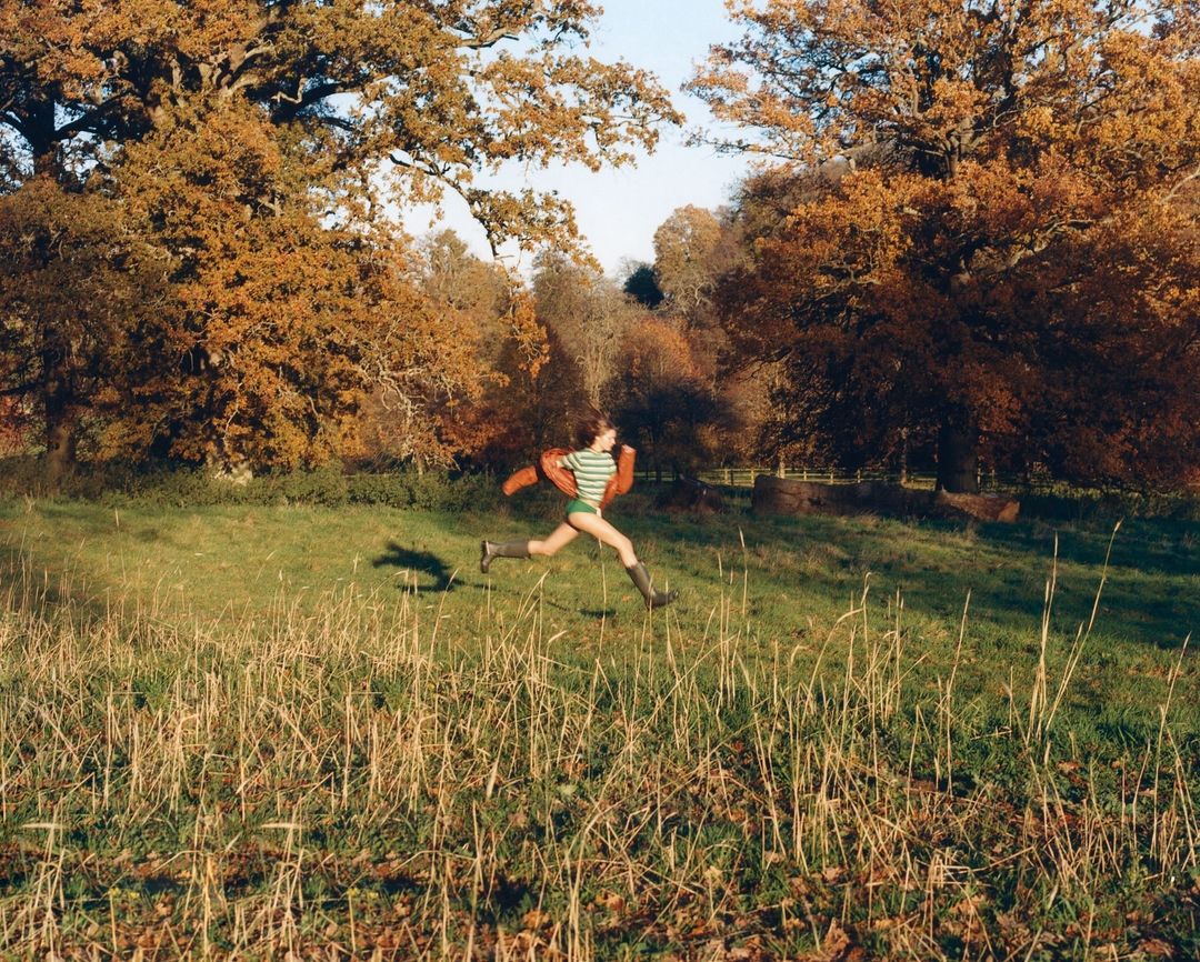 Woman in green swimsuit and jacket sprints through a grassy field in a forest with fall foliage, bathed in natural light.