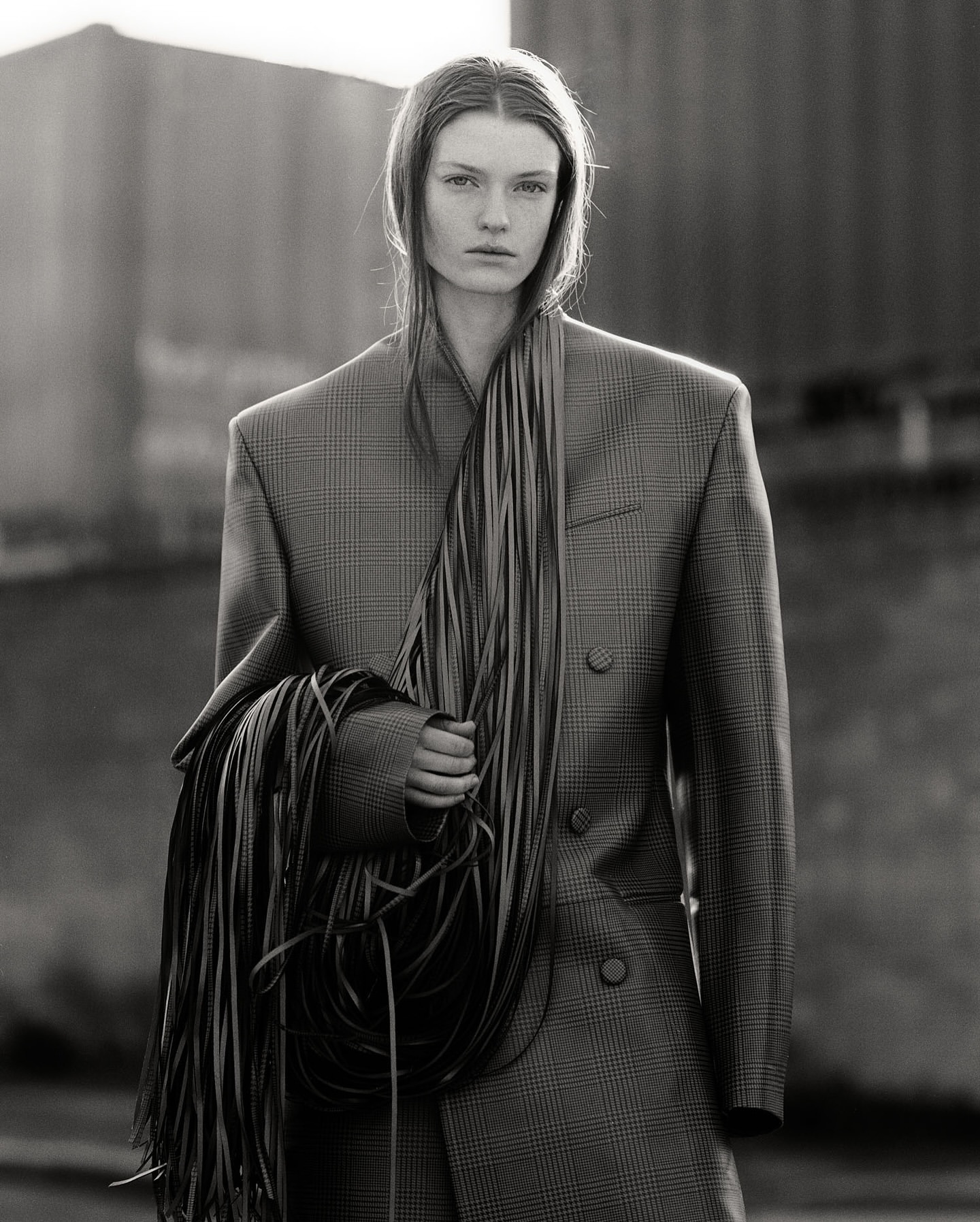 Monochrome fashion shot of a young adult female in a blazer holding a collection of cords, set against an out-of-focus urban background.