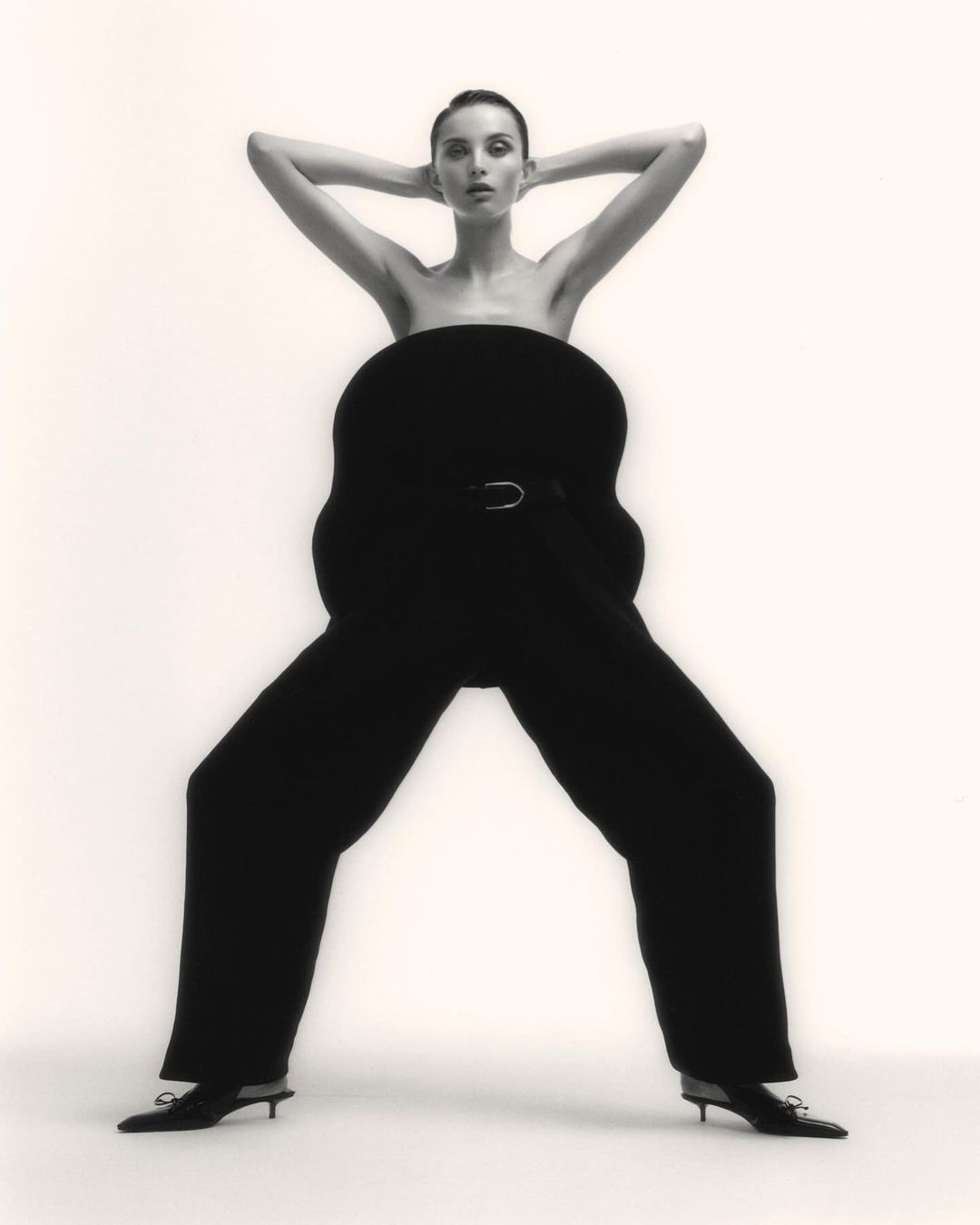 Woman poses in studio wearing oversized black pants and matching shoes. Her hands are behind her head, and she is facing forward.