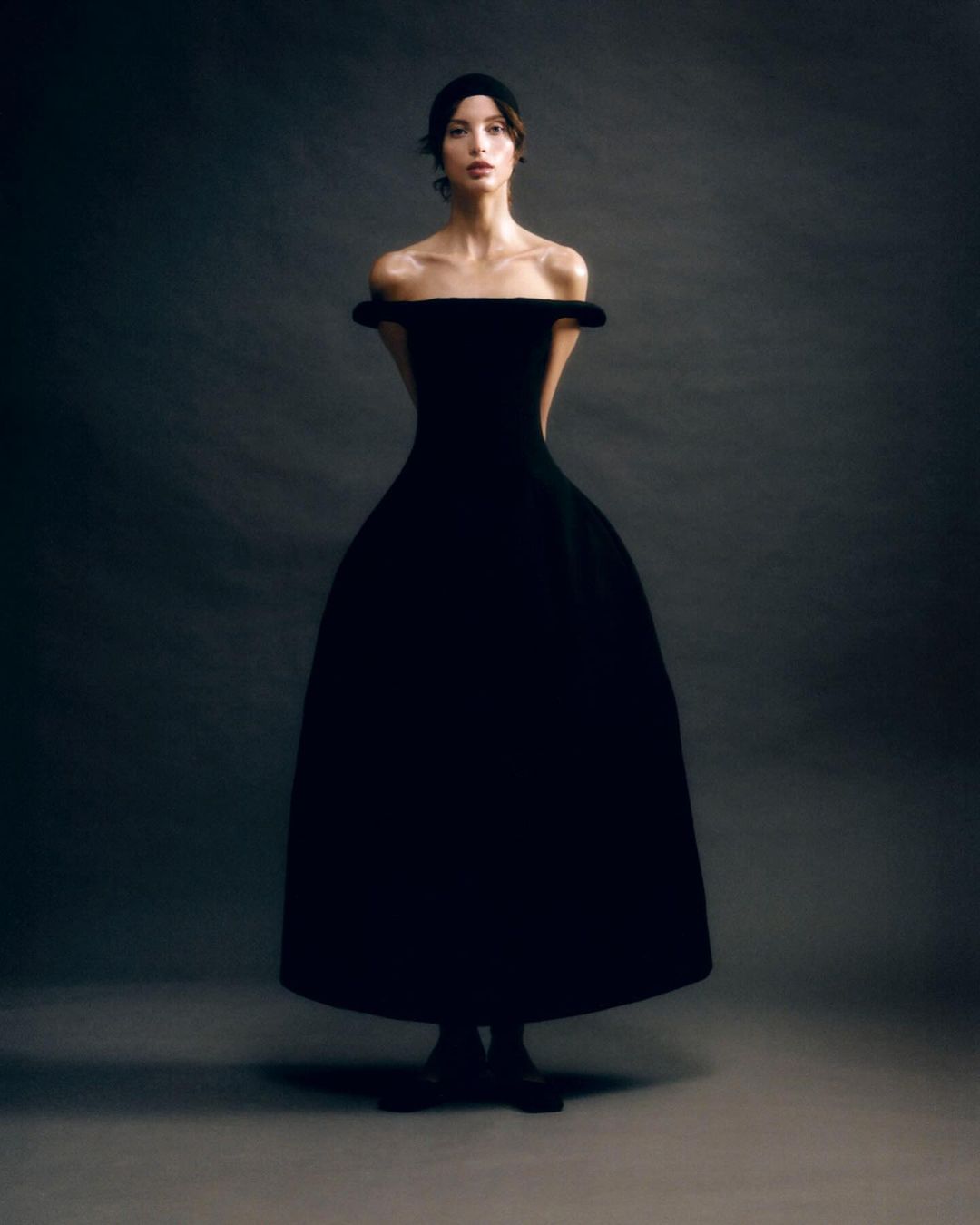 Woman in a black off-the-shoulder gown with headband poses in a photo studio with a simple gray background, shot in portrait orientation.