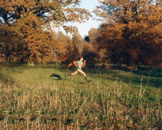 Woman in a green and white striped shirt and tall boots running through an autumn meadow framed by large oak trees