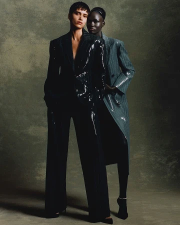 Two models standing against a mottled dark background, wearing a glossy black suit and a grey pinstripe coat.