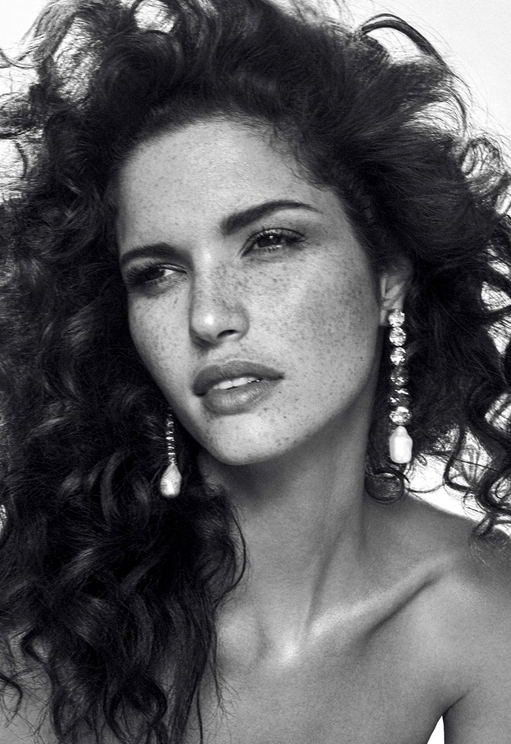 Black and white portrait of a woman with curly hair and freckles wearing dangling earrings against a bright, neutral studio backdrop.