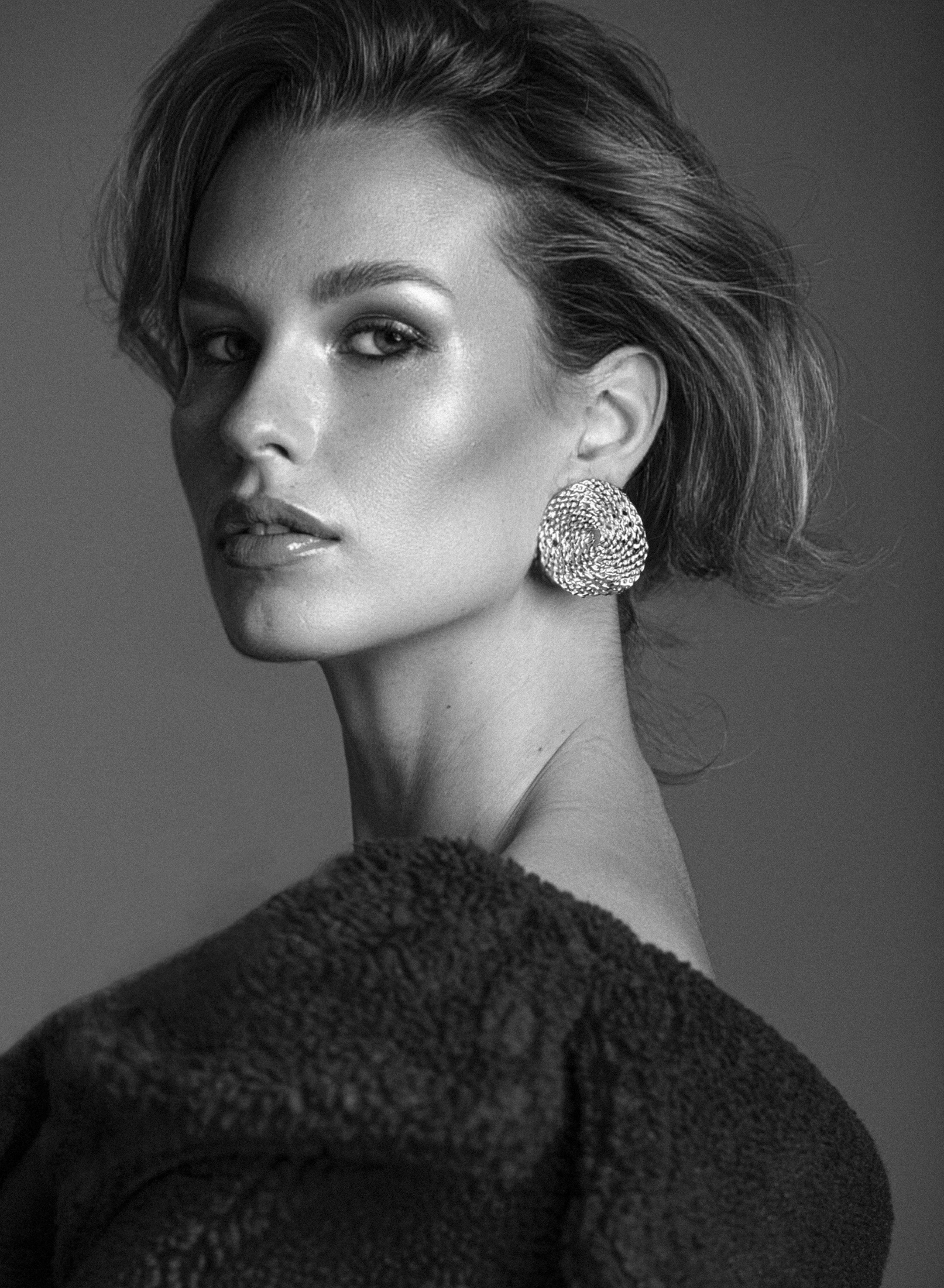 Black and white portrait of a woman wearing a statement earring and off-the-shoulder top, with a neutral gray background.
