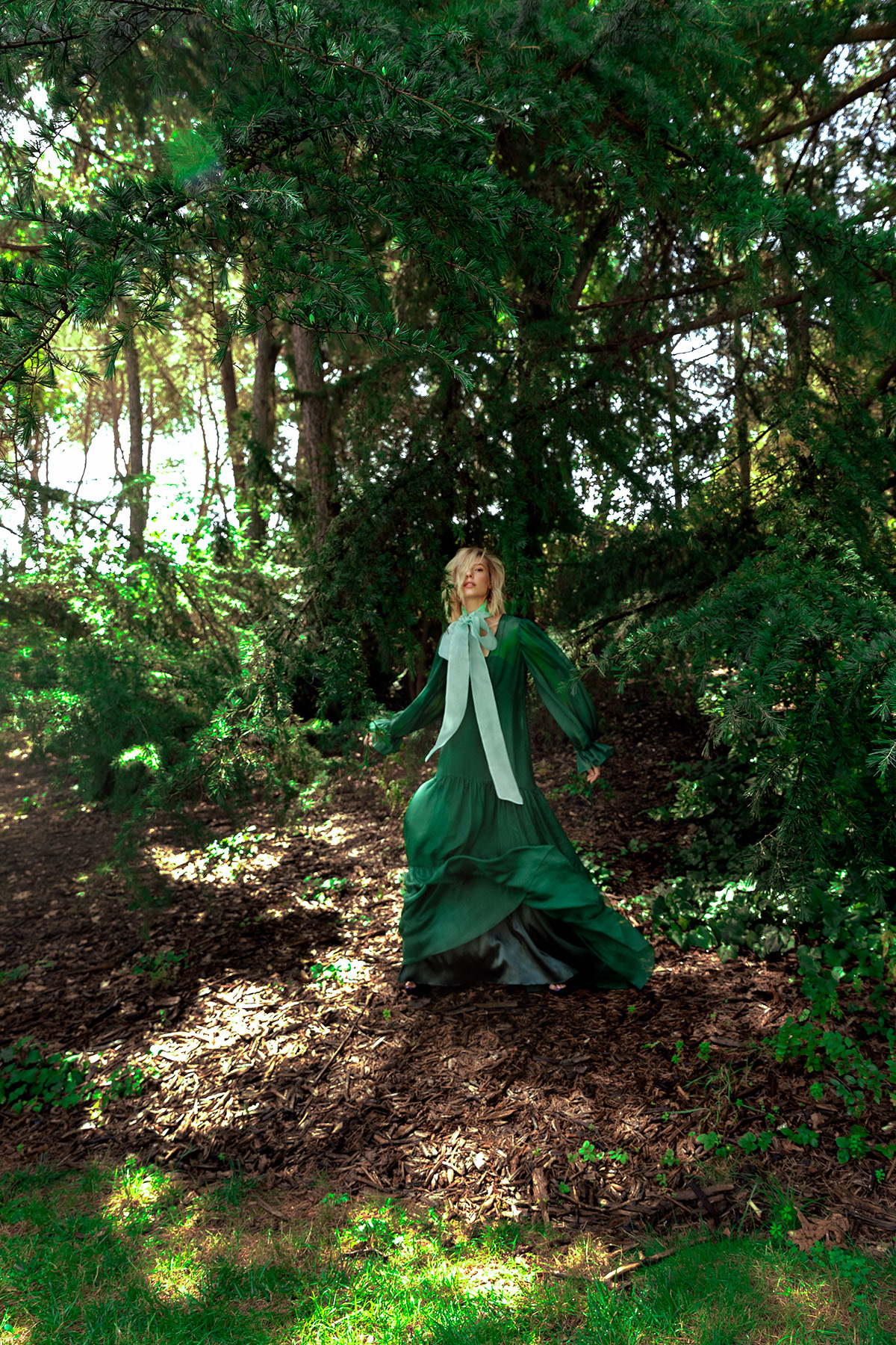 A blonde woman poses in a green dress and white bow in a wooded area with dappled light on the forest floor and lush trees.