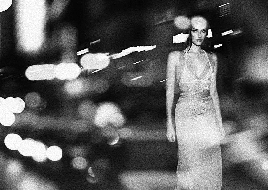 A woman in a formal dress stands in an urban environment at night, with blurred street lights adding depth to the black and white composition.