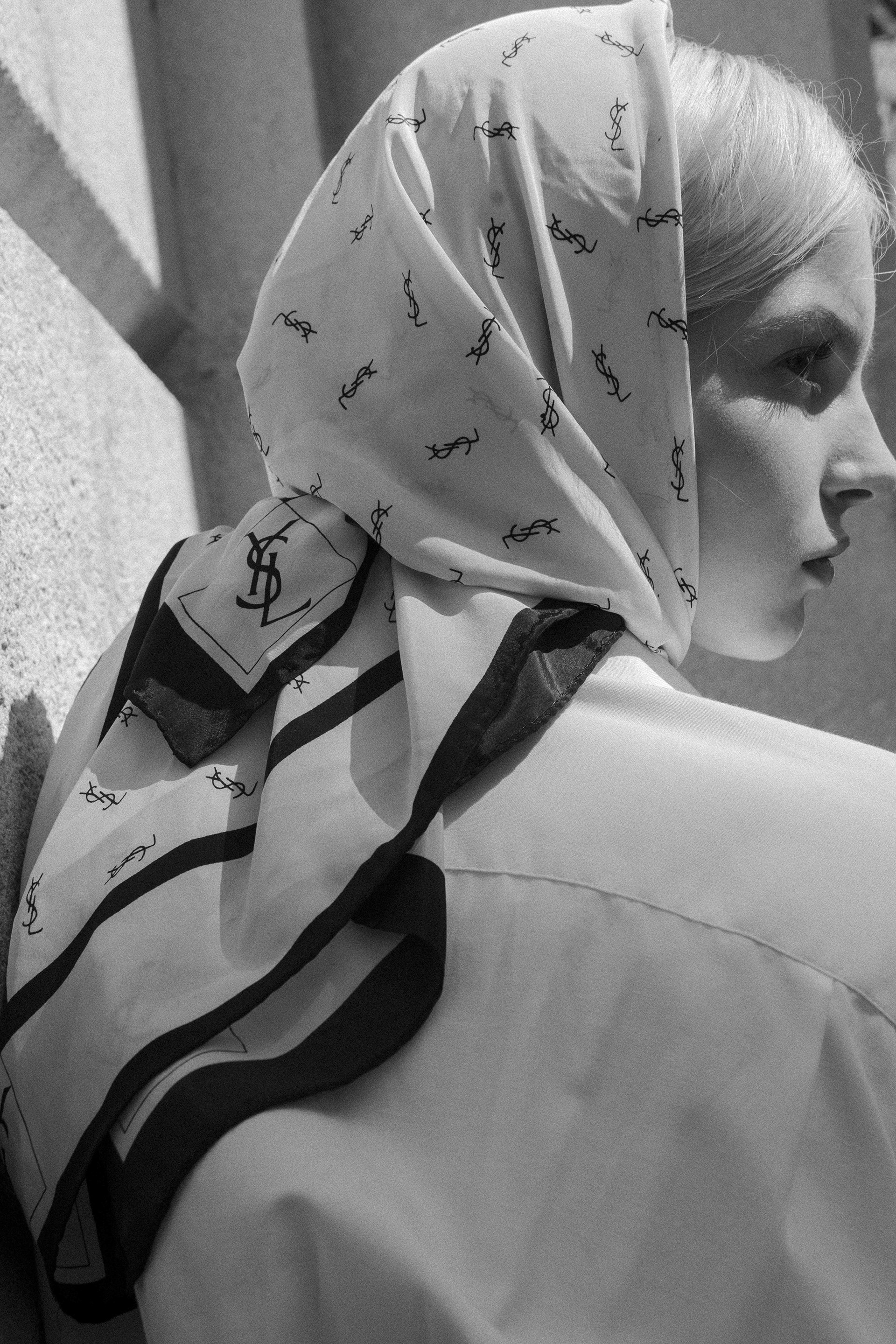 Woman in a white silk headscarf with black logo print, wearing a white collared shirt, posing in an outdoor setting with natural light.