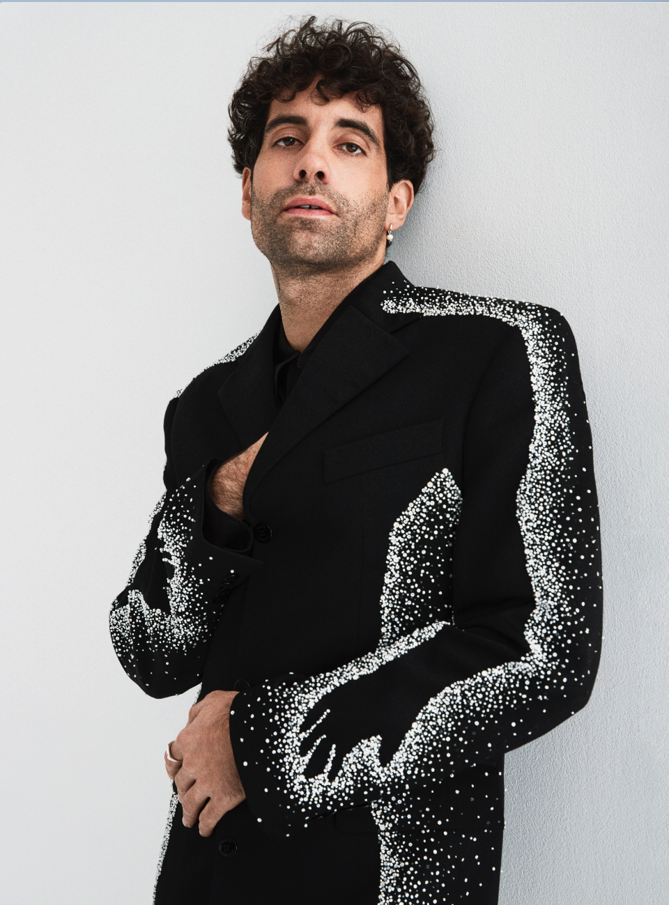 Headshot of a man with curly hair wearing a black blazer adorned with white beads, posing against a light grey wall, looking at the camera.