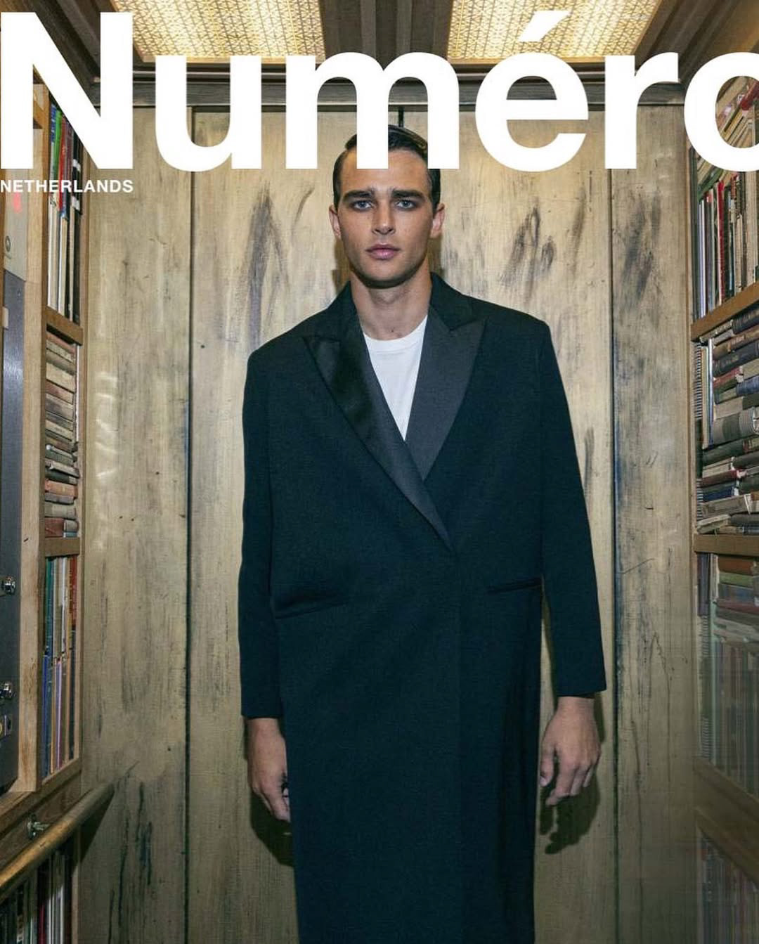 Numero magazine cover featuring an adult male model with dark hair in a black overcoat, standing in front of a wood-paneled backdrop with books.