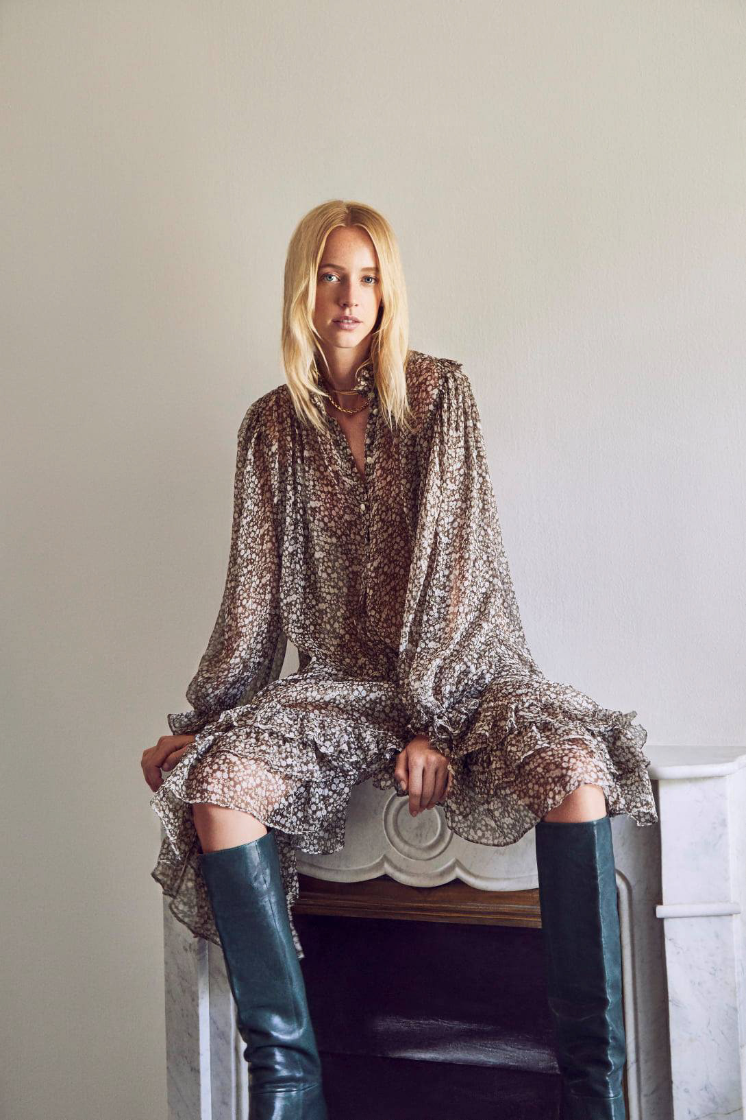 A blonde woman in a long-sleeve printed dress and tall boots sits on a marble fireplace mantel in a fashion-style indoor shot.