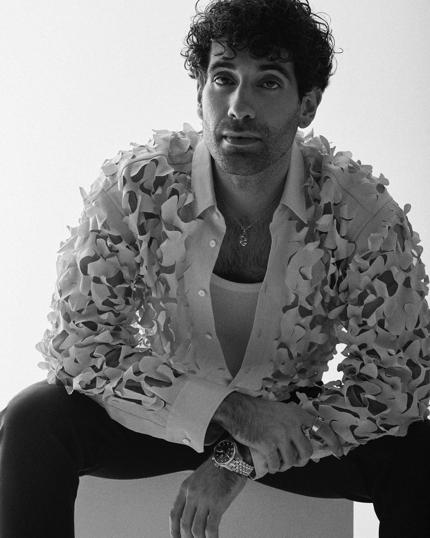 Monochrome portrait of a stylish adult male with curly dark hair, wearing a floral textured shirt, seated in front of a plain white background.