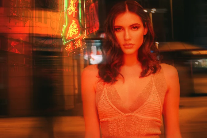 Adult female model with long hair poses in an orange-toned urban night scene with blurred neon signs and pedestrian movement behind her.