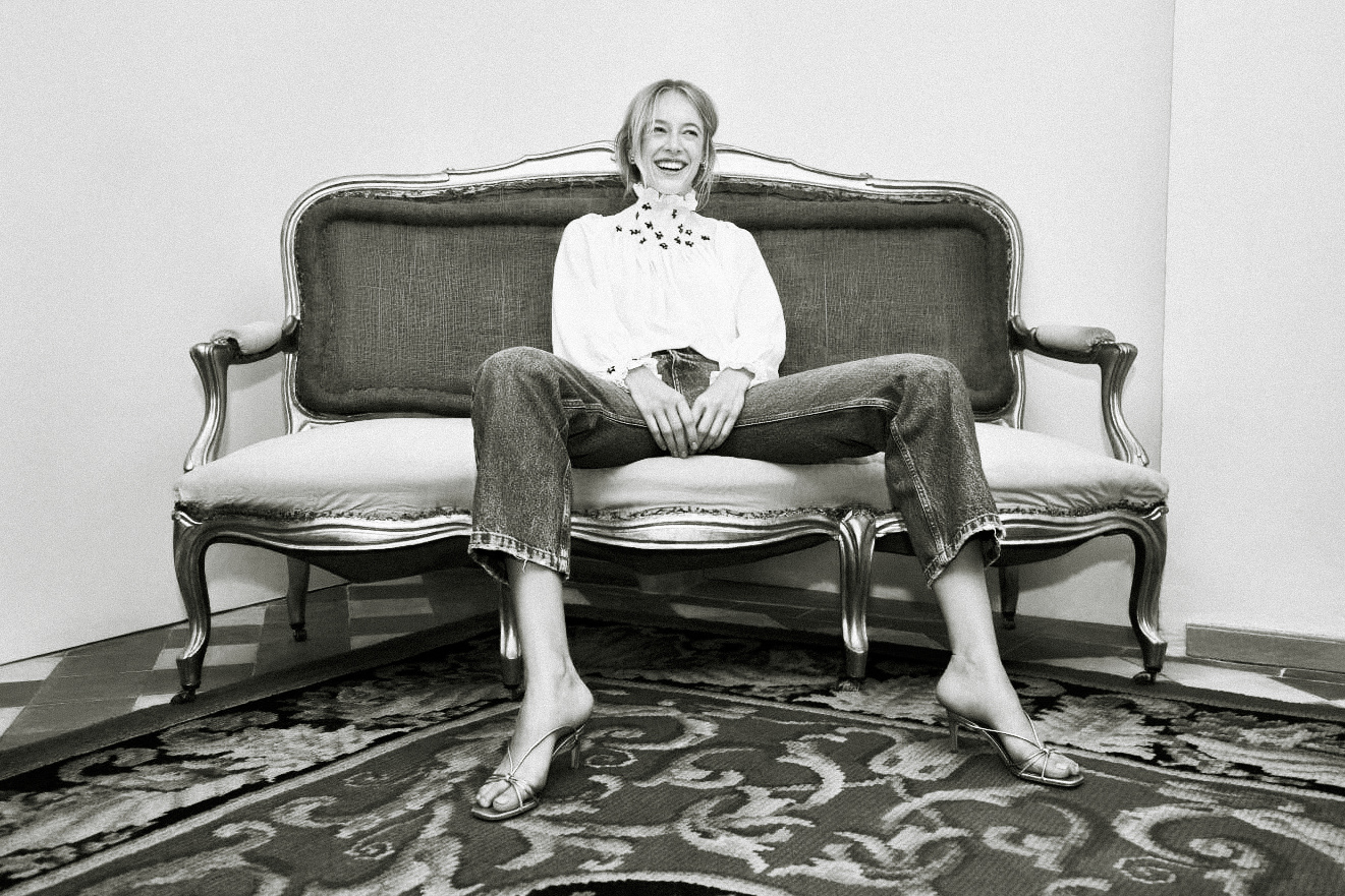 Blonde woman sits barefoot on an antique sofa wearing jeans and a white blouse in a studio setting with a decorative rug.