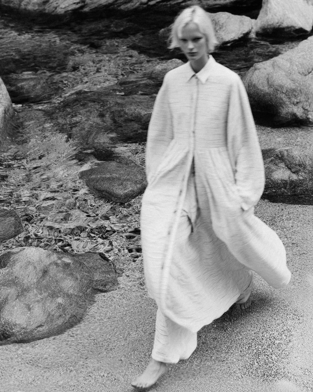 Monochrome fashion shot: a blonde adult woman walks barefoot through shallow water wearing a long, light-colored tunic and pants.