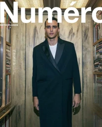 Man wearing a long dark coat and white t-shirt standing between wooden bookshelves in a library by Sera Zelva