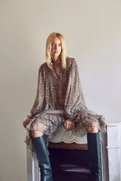 Blonde woman wearing a patterned ruffled dress and dark boots, seated on a mantelpiece in an interior setting.