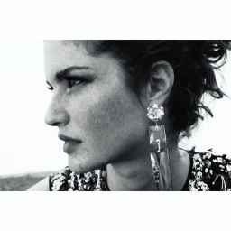 Close-up monochrome portrait of a woman wearing large crystal drop earrings and a sequined top against an outdoor sky