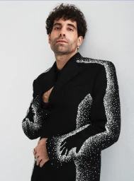 Man with curly hair wearing a black jacket with rhinestone embellishments against a plain light wall.