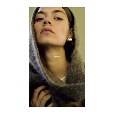 Woman with brown hair wearing chunky gold earrings and a grey sweater in a close-up fashion studio portrait.