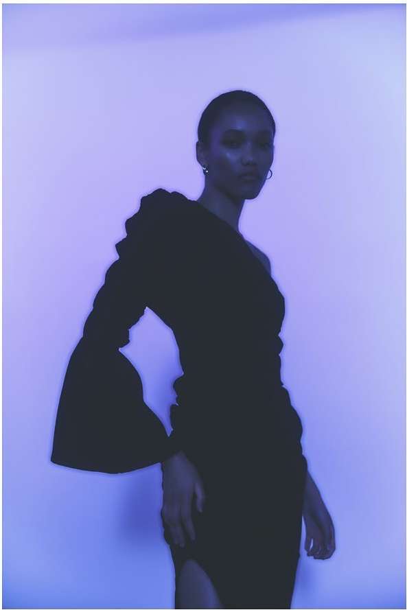 Elegant Black woman in a one-shoulder black dress with flared sleeve posing against a soft purple backdrop in a studio setting.