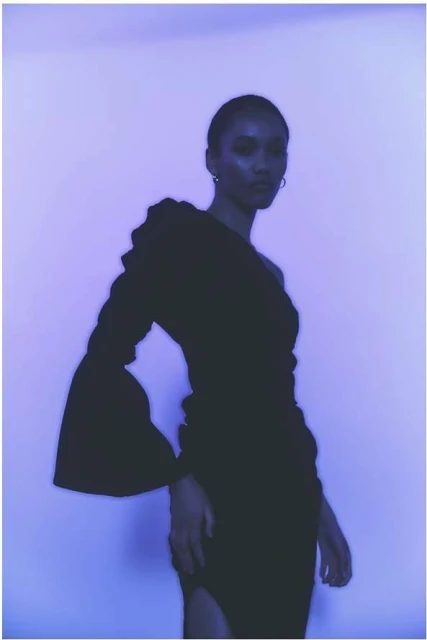 Elegant Black woman in a one-shoulder black dress with flared sleeve posing against a soft purple backdrop in a studio setting. 