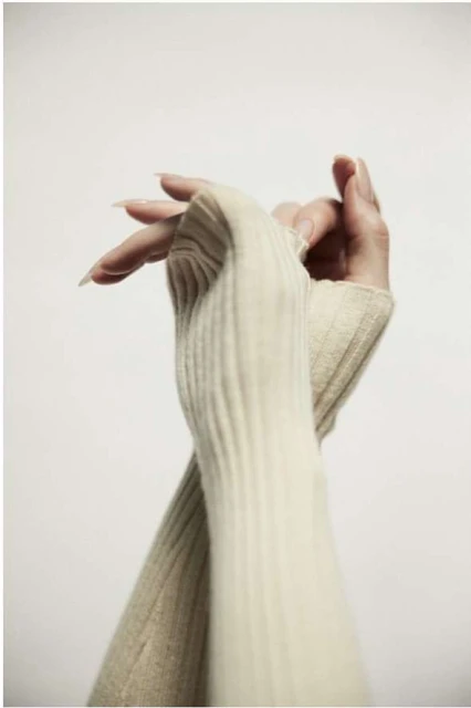 Close-up of two adult female hands crossed in front of a neutral background, wearing cream-colored, ribbed, fingerless knit gloves.